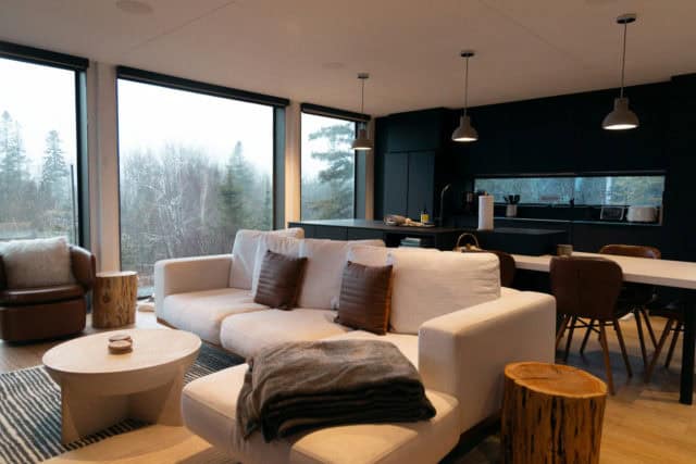 Photo d'un intérieur moderne prise depuis le salon et en direction d'une cuisine ouverte à dominante de noir. Il y a de grande baie vitrée laissant passer beaucoup de lumière - ArchiBienPensé, architecte d'intérieur à Annecy et en Haut-Savoie