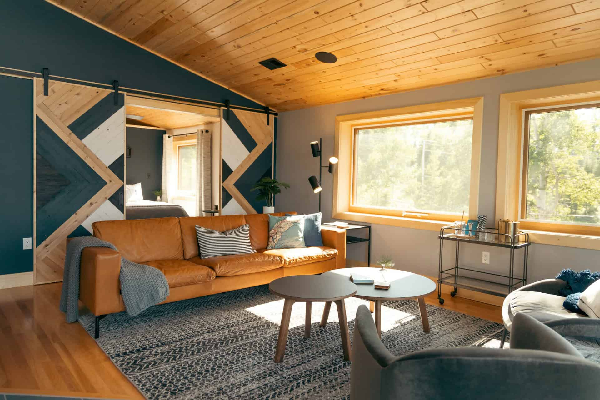 Photo de l'intérieur moderne et ensoleillé d'un chalet en montagne, avec des prédominantes de couleurs bleu foncé désaturé, bois et marron. Il y a un canapé, deux tables centrales et de belles grande fenêtres