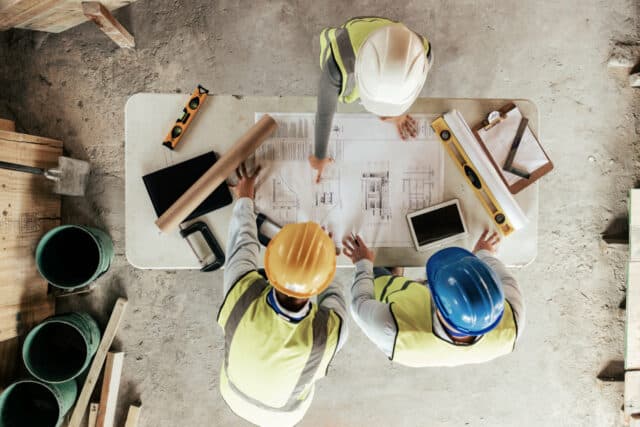 Trois artisans vus de dessus sont en train de consulter les plans d'un chantier pour coordonner les travaux - Maîtrise d'œuvre et gestion de chantier à Annecy et en Haute-Savoie