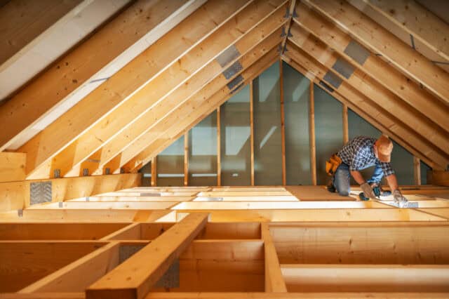 Un charpentier mesure et ajuste soigneusement des poutres en bois dans un grenier, éclairé par la lumière du soleil filtrant à travers le toit. L'espace de travail reflète un effort de rénovation - Maîtrise d'œuvre et gestion de chantier à Annecy et en Haute-Savoie