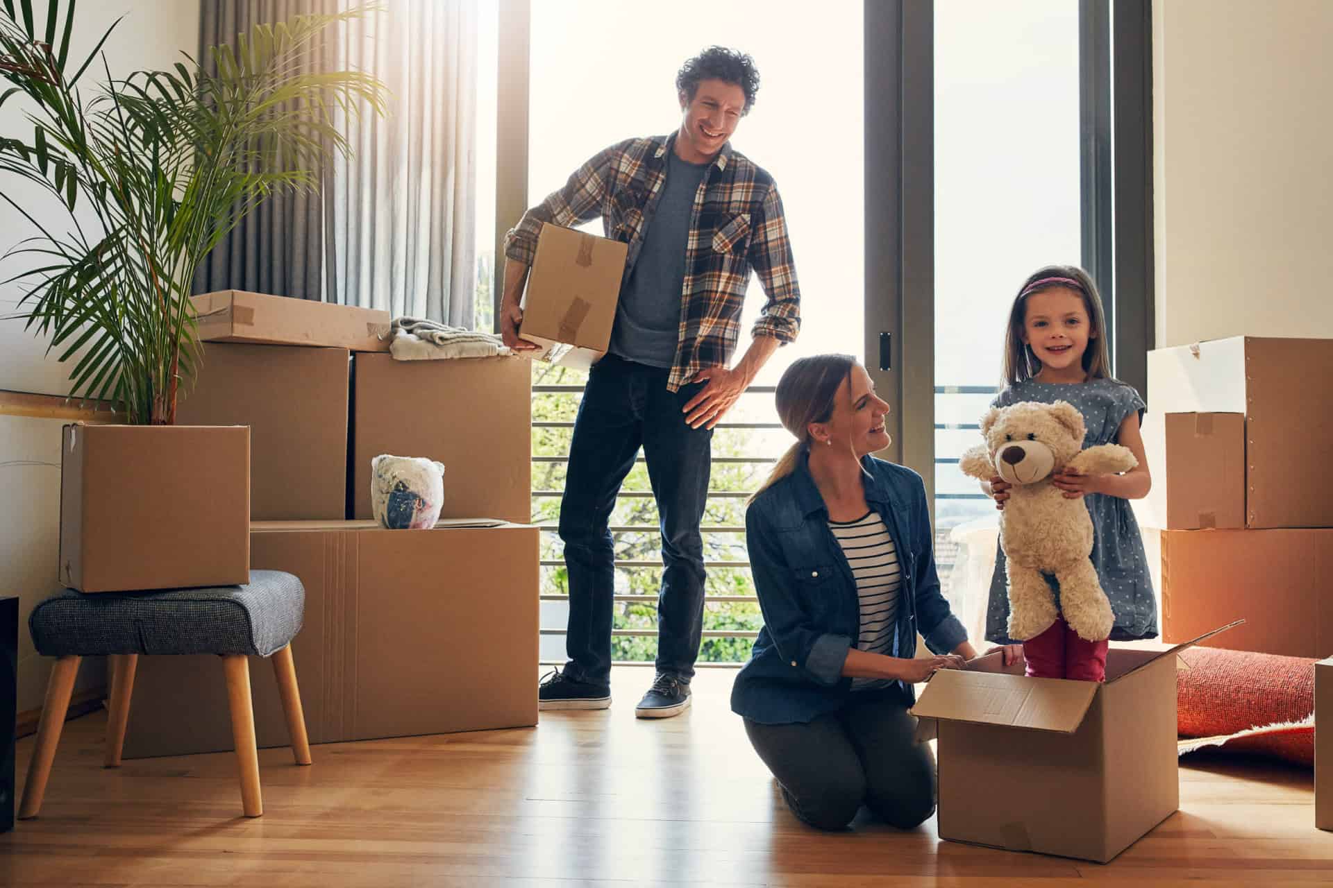 Une famille de trois personnes déballe les cartons pour s'installer dans leur nouvel intérieur. Une petite fille tient un ours en peluche