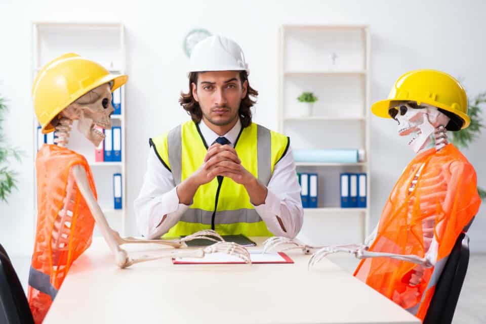 Un homme portant un casque et un gilet de sécurité, assis à une table avec deux squelettes portant des gilets et des casques également.